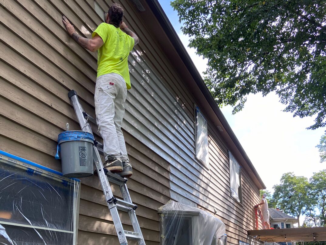 man in yellow shirt standing tall on a ladder painting a house