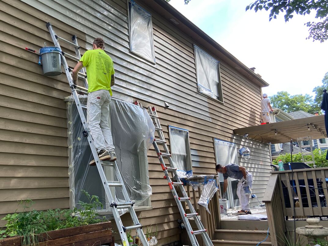 three men working in the sun to paint the side of a house